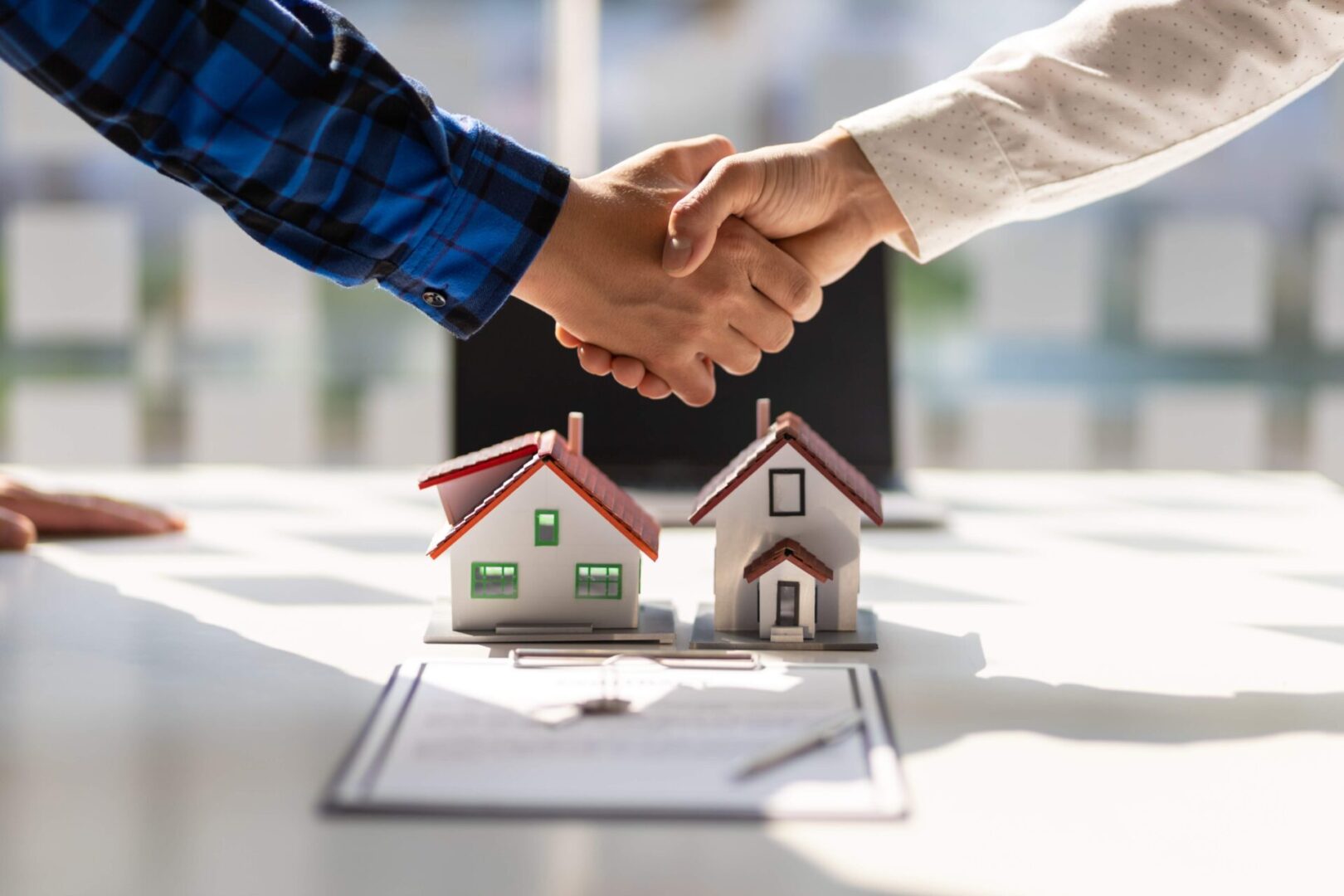 Two people shaking hands over miniature houses and a contract, symbolizing a real estate deal.